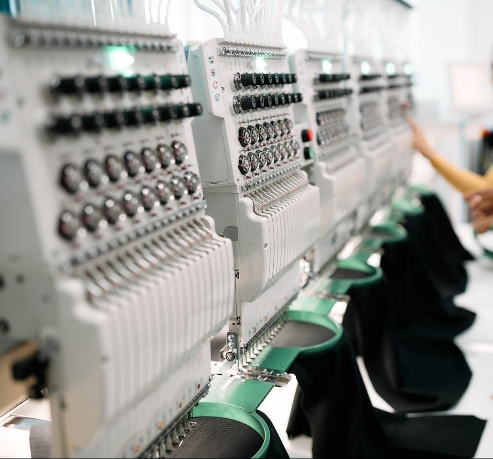 Close up of embroidery machine in textile factory. Women are working in the background.
